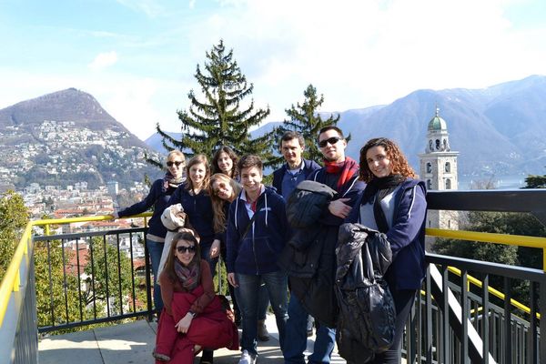 Nove ragazzi e ragazze su una terrazza con la ringhiera gialla; alle spalle la città sulle pendici di un monte e un campanile con la cupola verde
