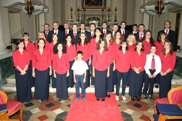 Foto di gruppo davanti a un altare con sei candelieri: donne in camicia rossa e gonna blu, uomini in abito scuro e cravatta rossa, in mezzo un bambino
