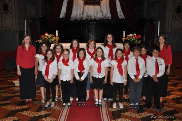 Una quindicina di bambini e bambine in camicia bianca, con foulard o cravatta rossa, tra due giovani donne in camicia rossa davanti all’altare
