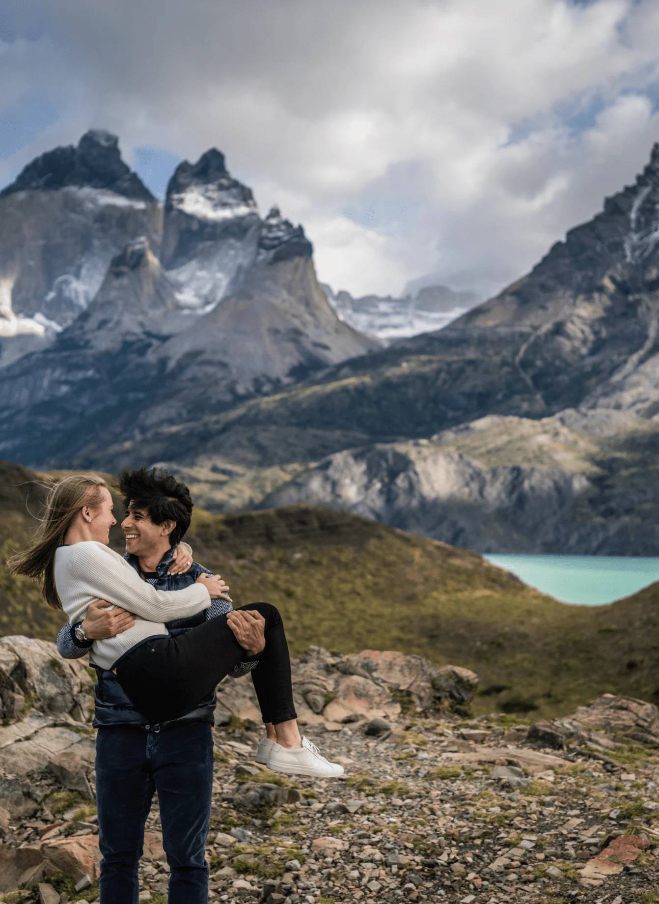 A man carrying his partner in his arms, laughing, with the Cuernos del Paine and a turquoise lake behind them