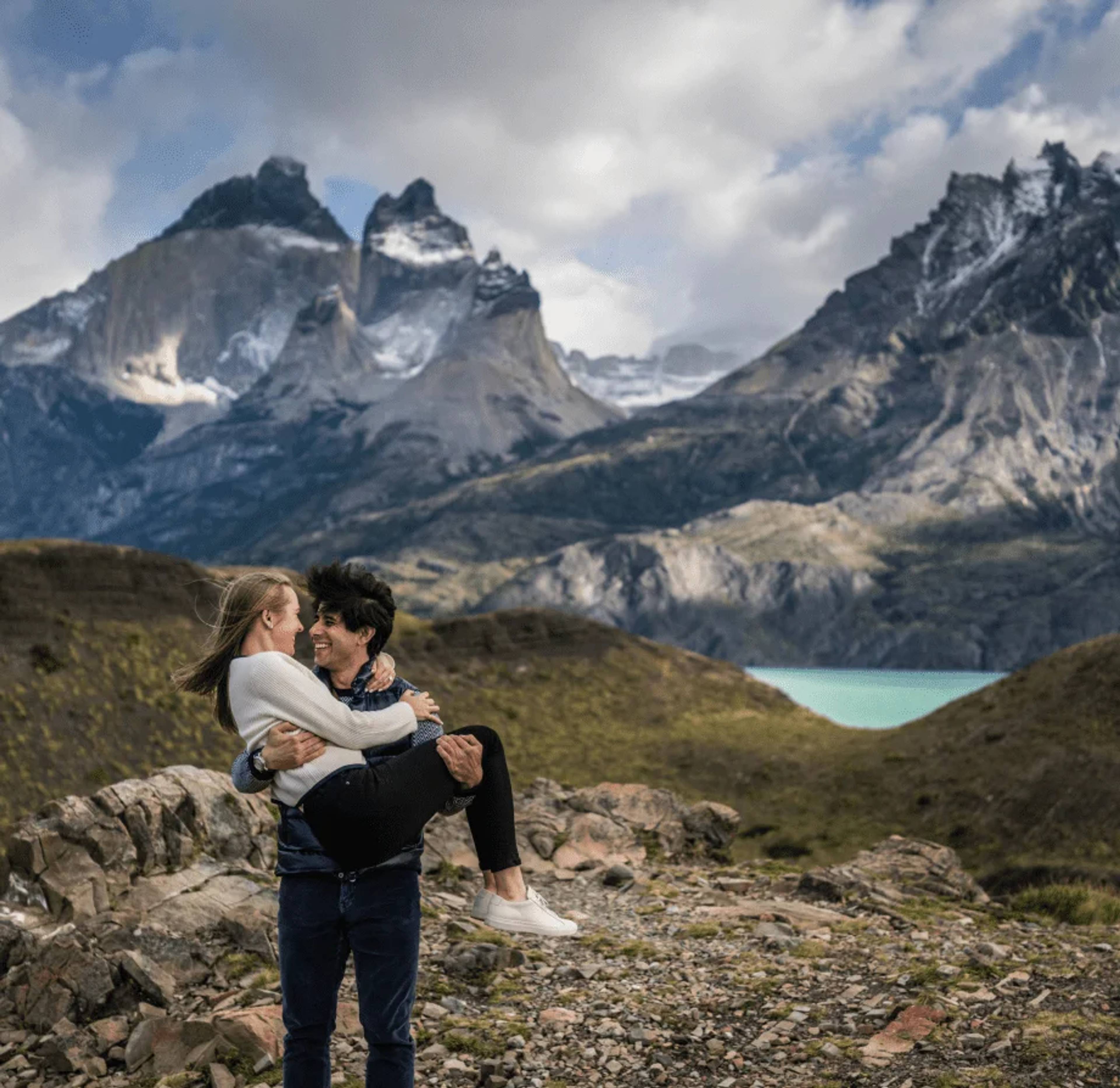 A couple laughing as he lifts her, the Cuernos del Paine wrapped in cloud behind them
