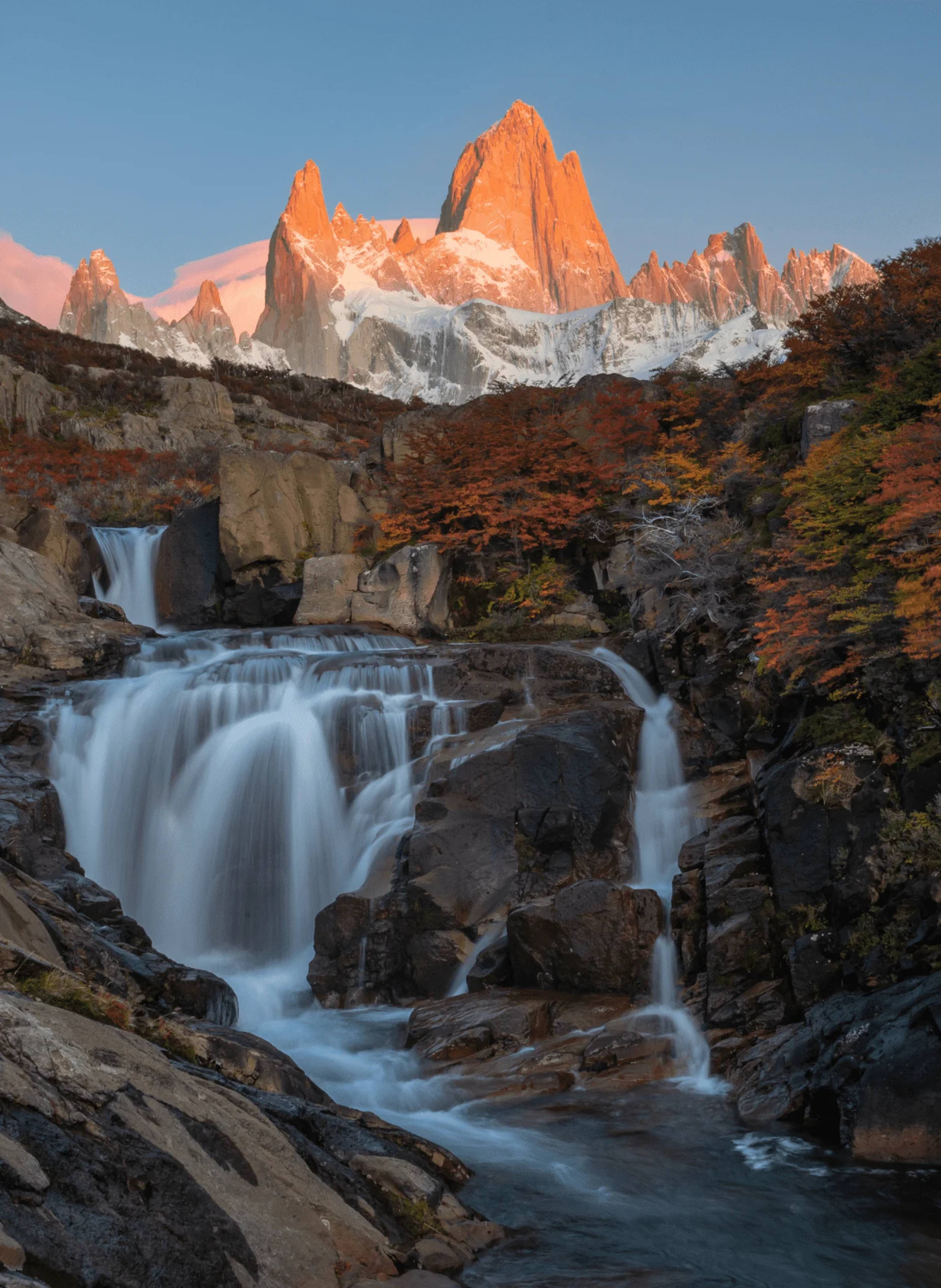 A waterfall in the foreground and the Fitz Roy massif lit pink at dawn, framed by autumn lenga trees