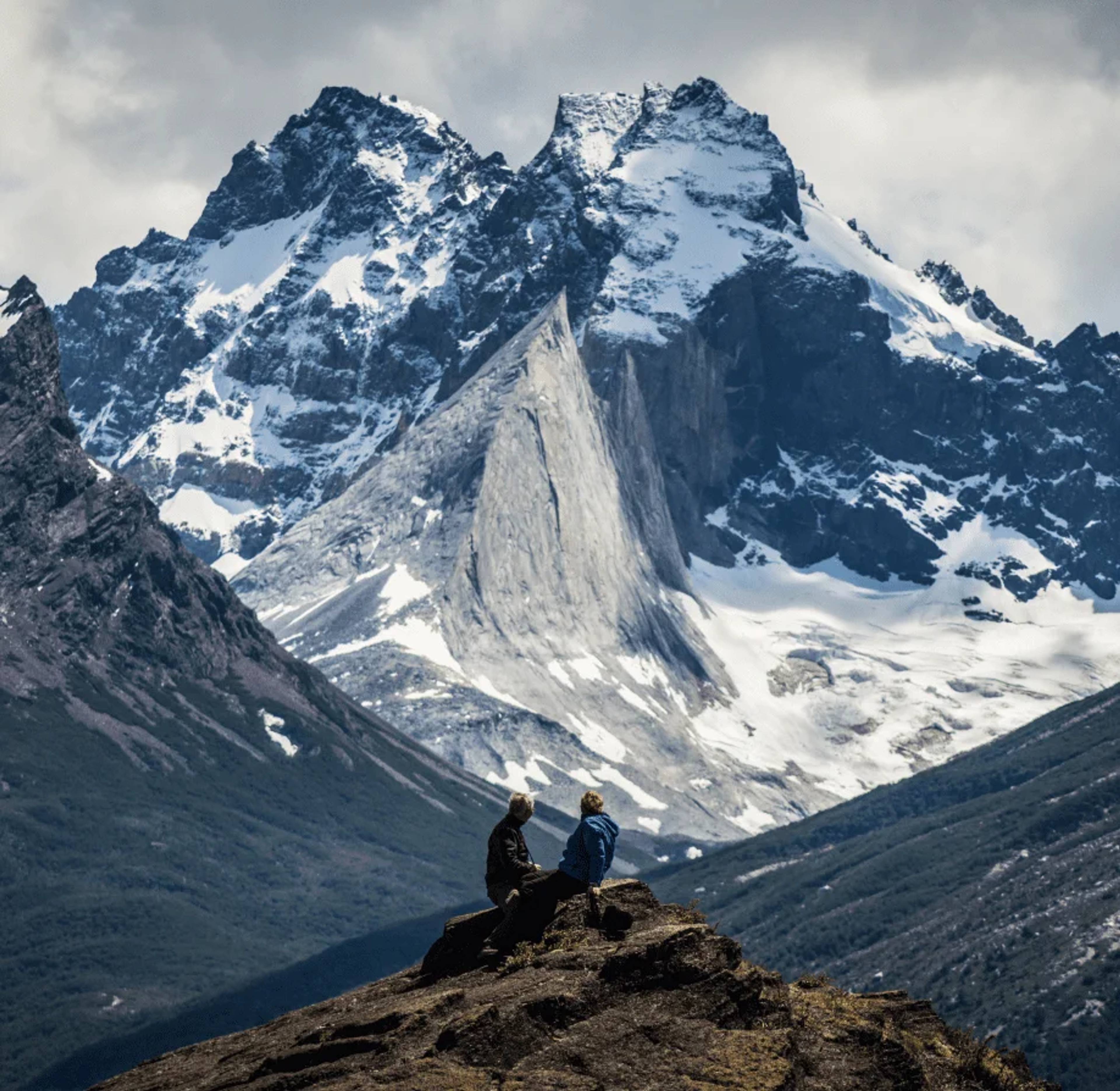 Two people sitting on a ridge looking out at a snow-covered peak across the valley