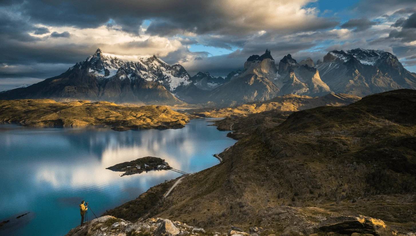 A lone photographer on a ridge above Lake Pehoé, dwarfed by the Paine massif under a broken sky