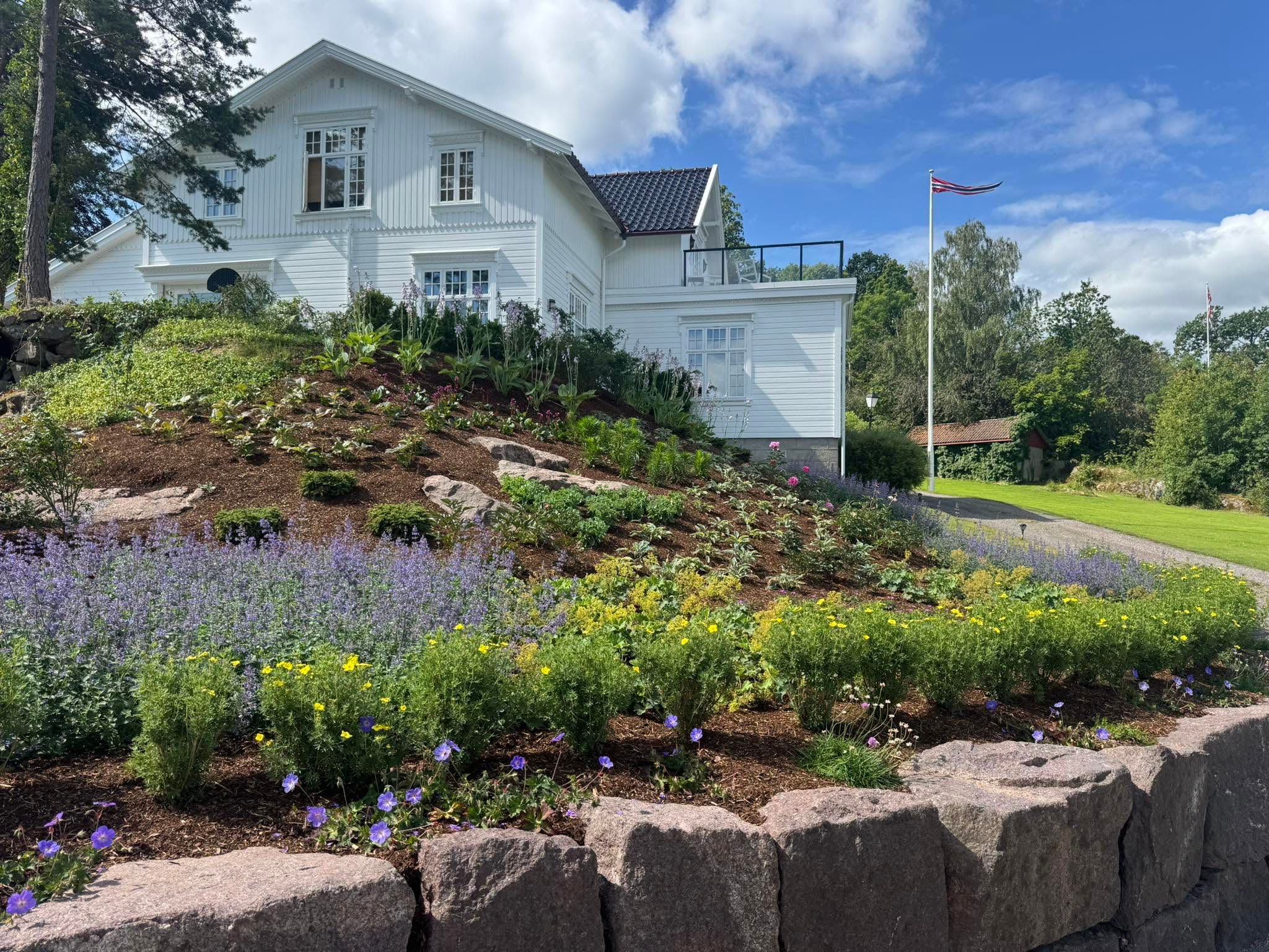 Støttemur i naturstein med frodig staudebed foran et hvitt hus