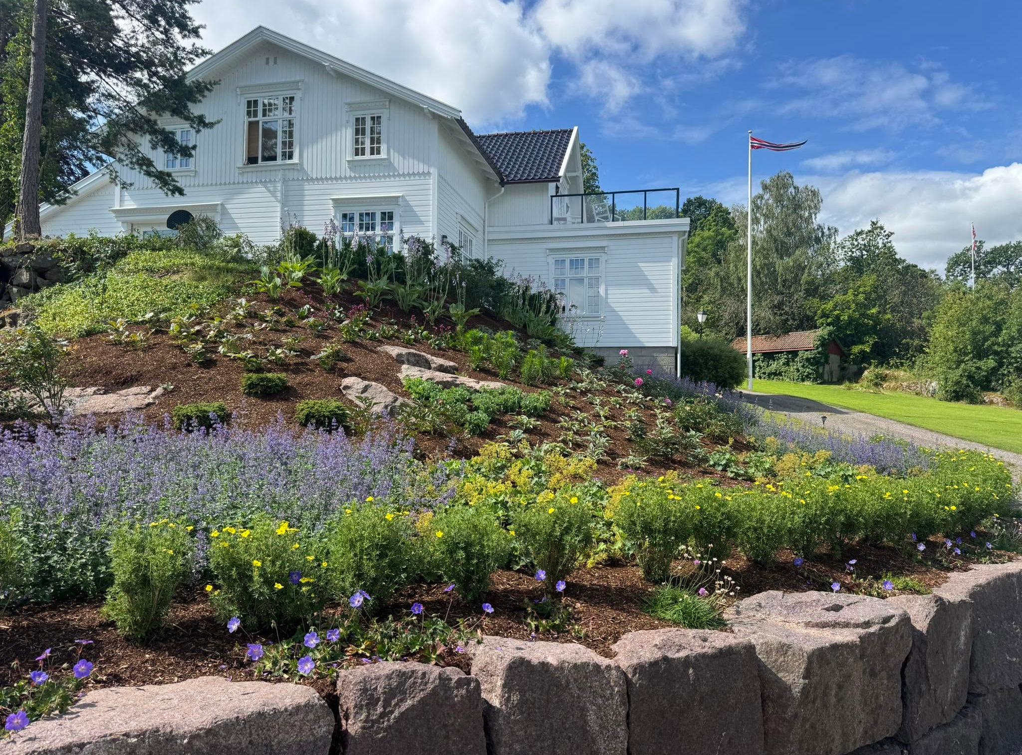Støttemur i naturstein med frodig staudebed foran et hvitt hus