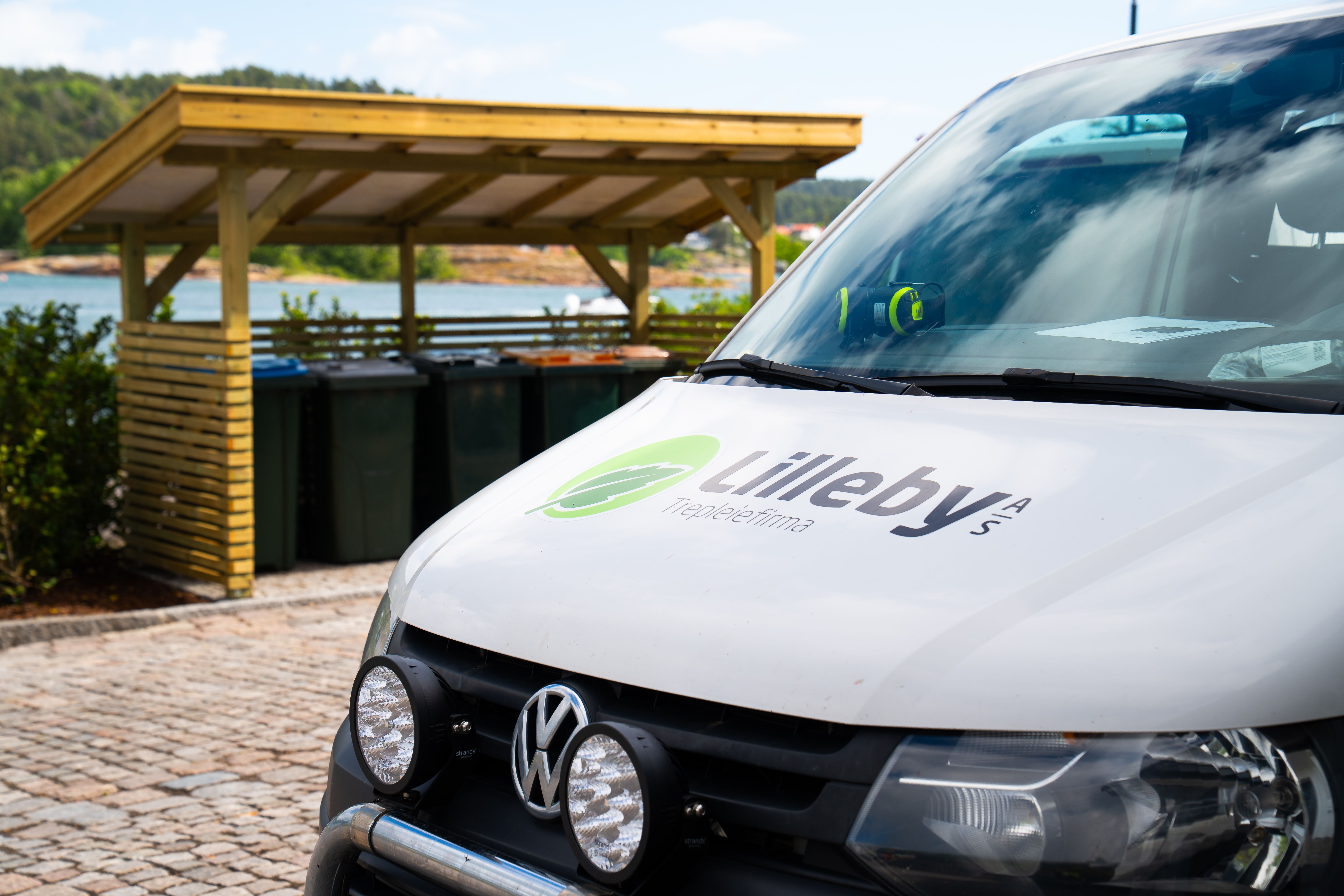 Hvit firmabil med Lilleby-logo parkert på brostein ved avfallsskuret