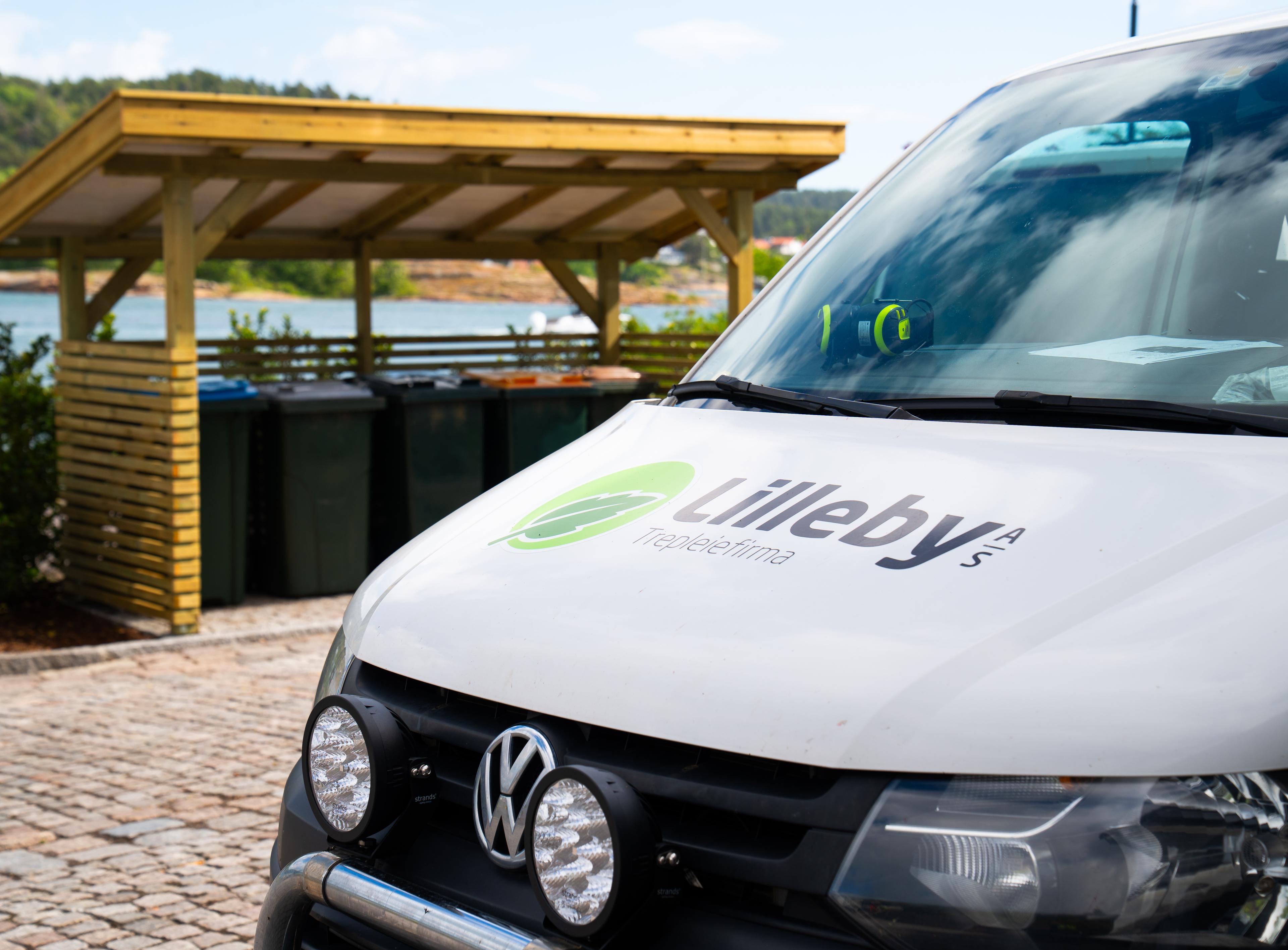 Hvit firmabil med Lilleby-logo parkert på brostein ved avfallsskuret
