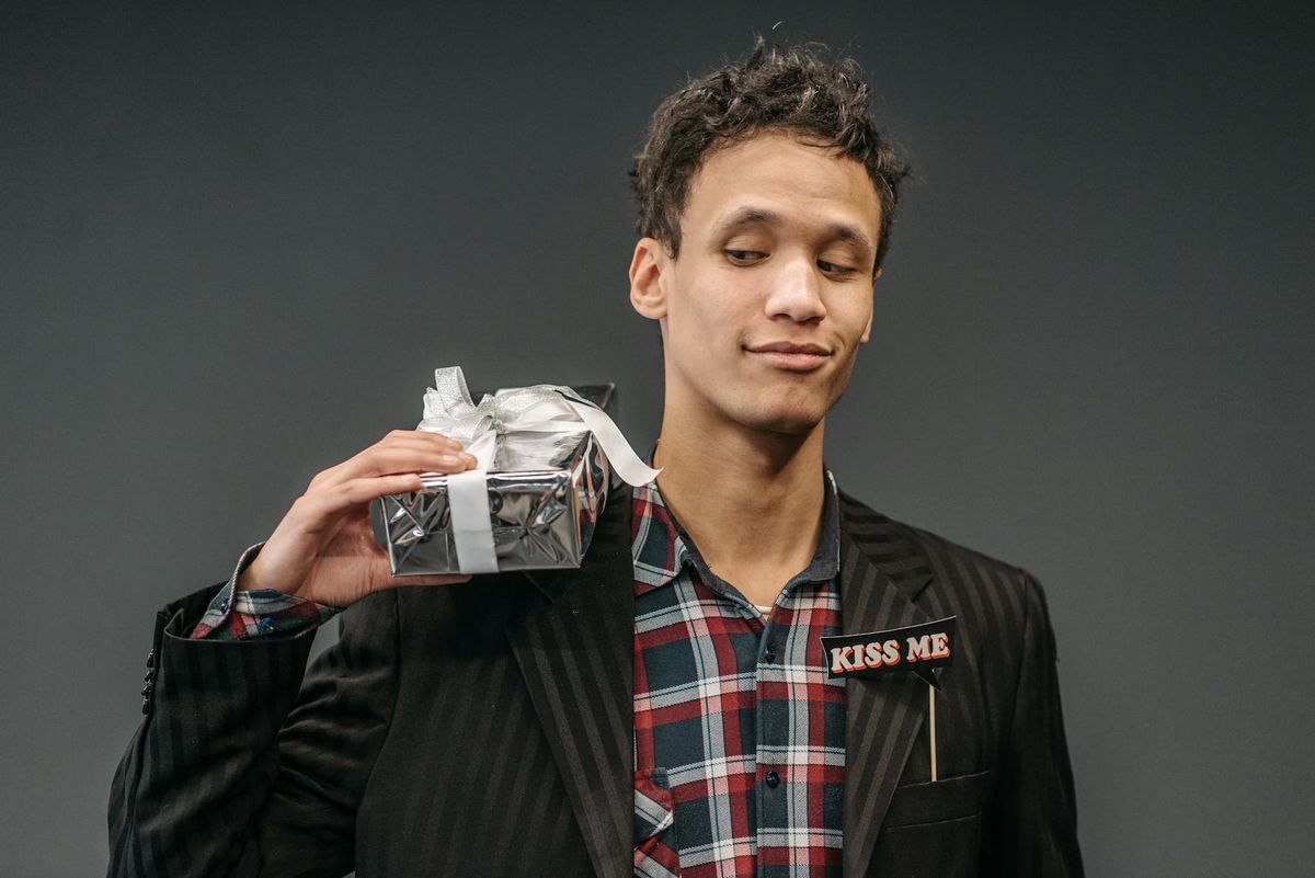 Young man in a plaid shirt and striped suit holds a gift, showcasing a playful expression in a studio setting.