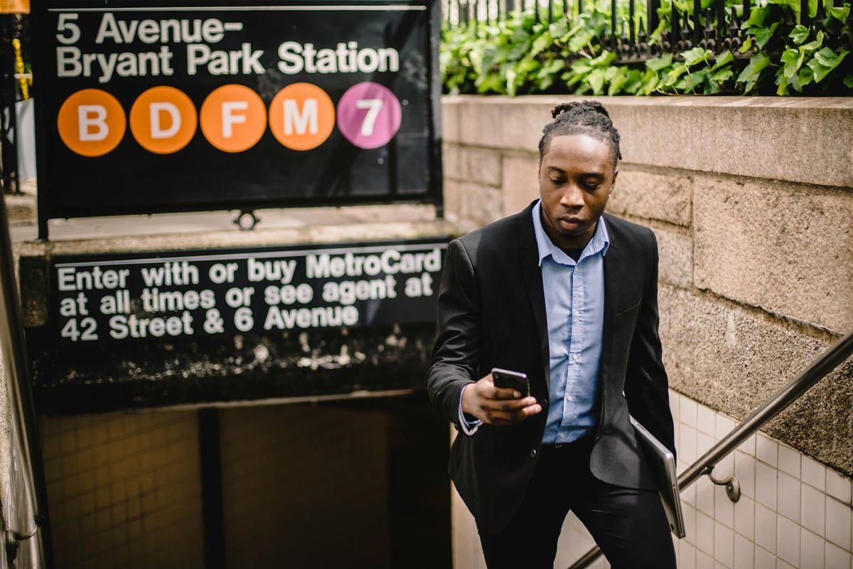 Serious African American office worker with laptop in hand using mobile while exiting subway in Manhattan on sunny summer day