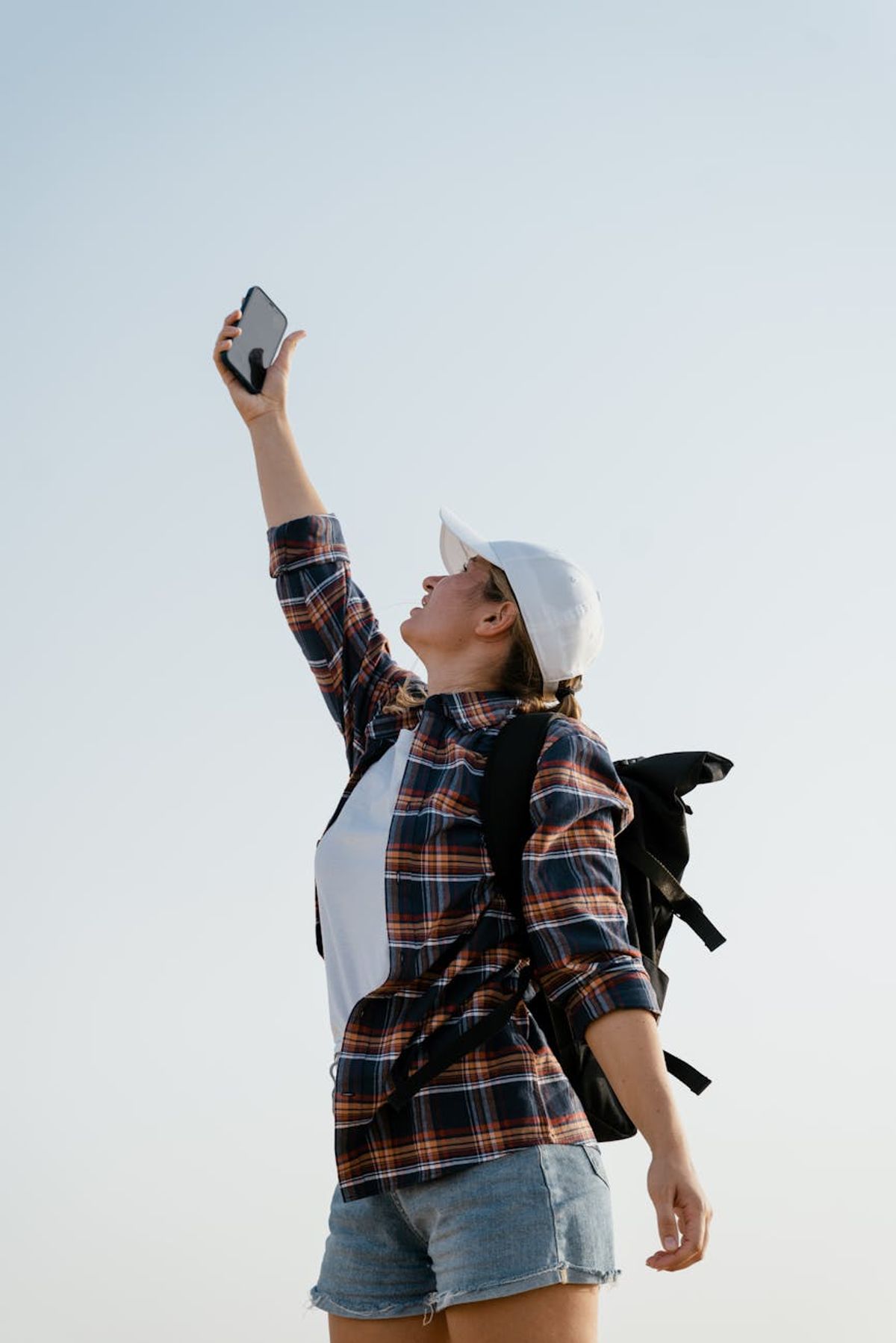 A young woman in casual attire reaches up with her smartphone outdoors for better signal.