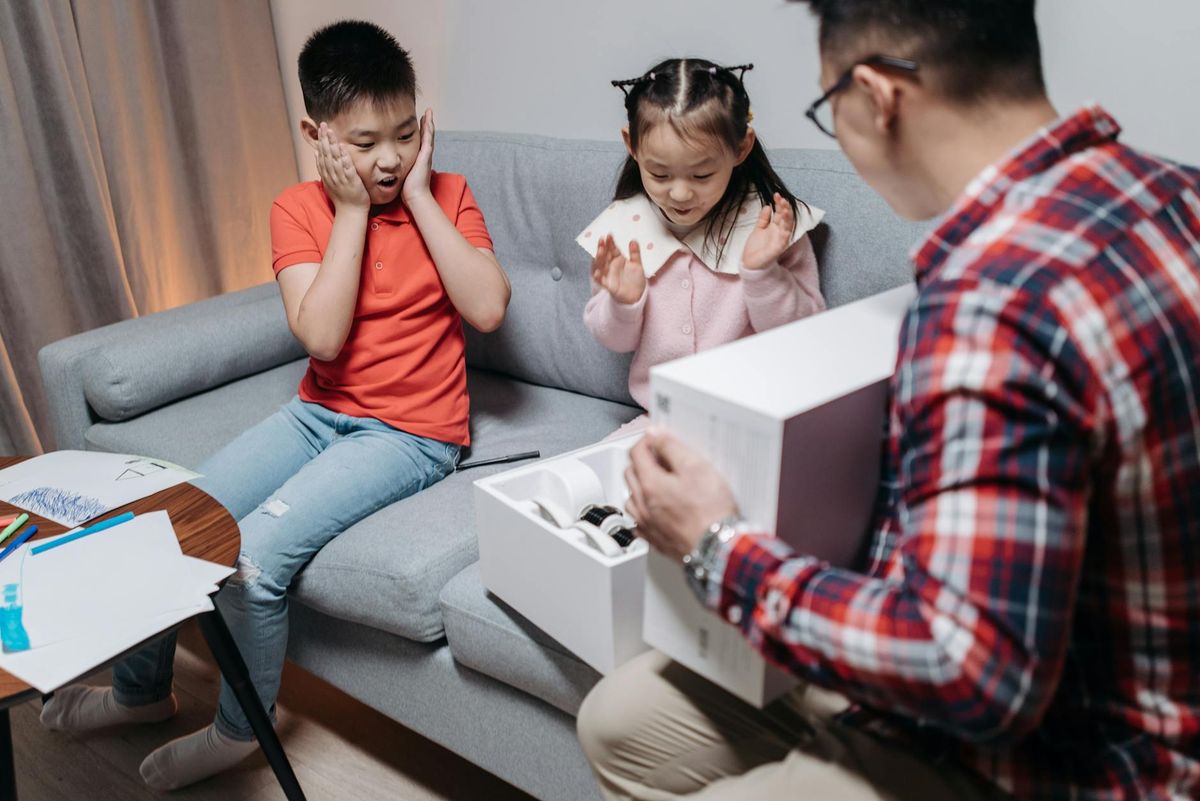Children on couch look amazed as father reveals a technology gift from a box.