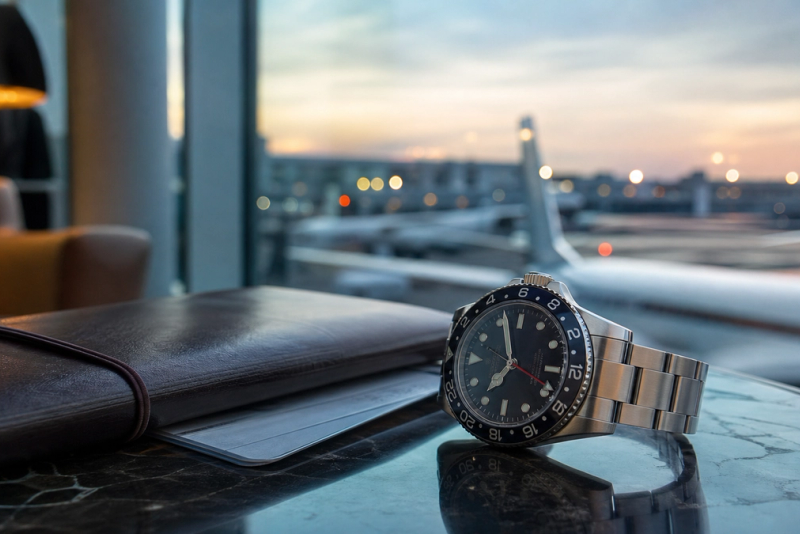 GMT-style mechanical watch beside a travel notebook in an airport lounge at dawn