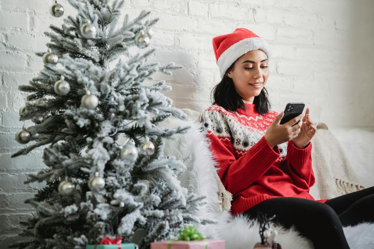 Happy young female in Santa hat and red jumper sitting on sofa near decorated Christmas tree and browsing mobile phone