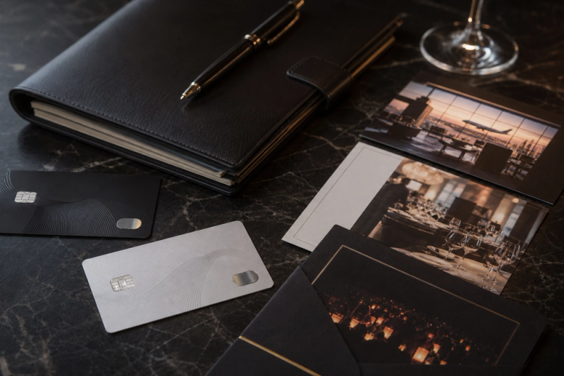 Two distinct unbranded premium payment cards beside a private itinerary folio, airport lounge and curated event invitation