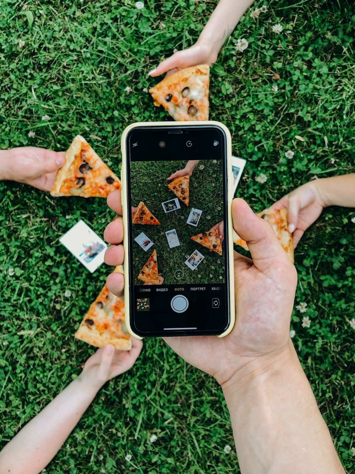 Top view of a picnic setting with hands holding pizza slices captured on a smartphone camera.