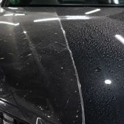 Macro editorial photograph of a dark luxury-car bonnet divided into two real protection demonstrations: transparent film edge absorbing small road debris on one side and water beading on a coated surface on the other