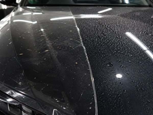 Macro editorial photograph of a dark luxury-car bonnet divided into two real protection demonstrations: transparent film edge absorbing small road debris on one side and water beading on a coated surface on the other
