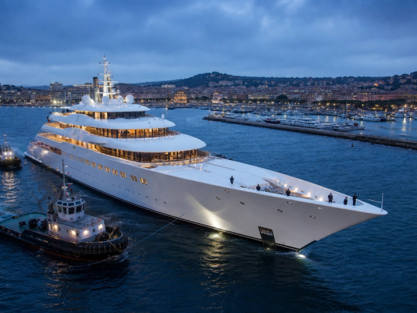 A record-scale white superyacht entering harbour with tugboats and crew visible for scale