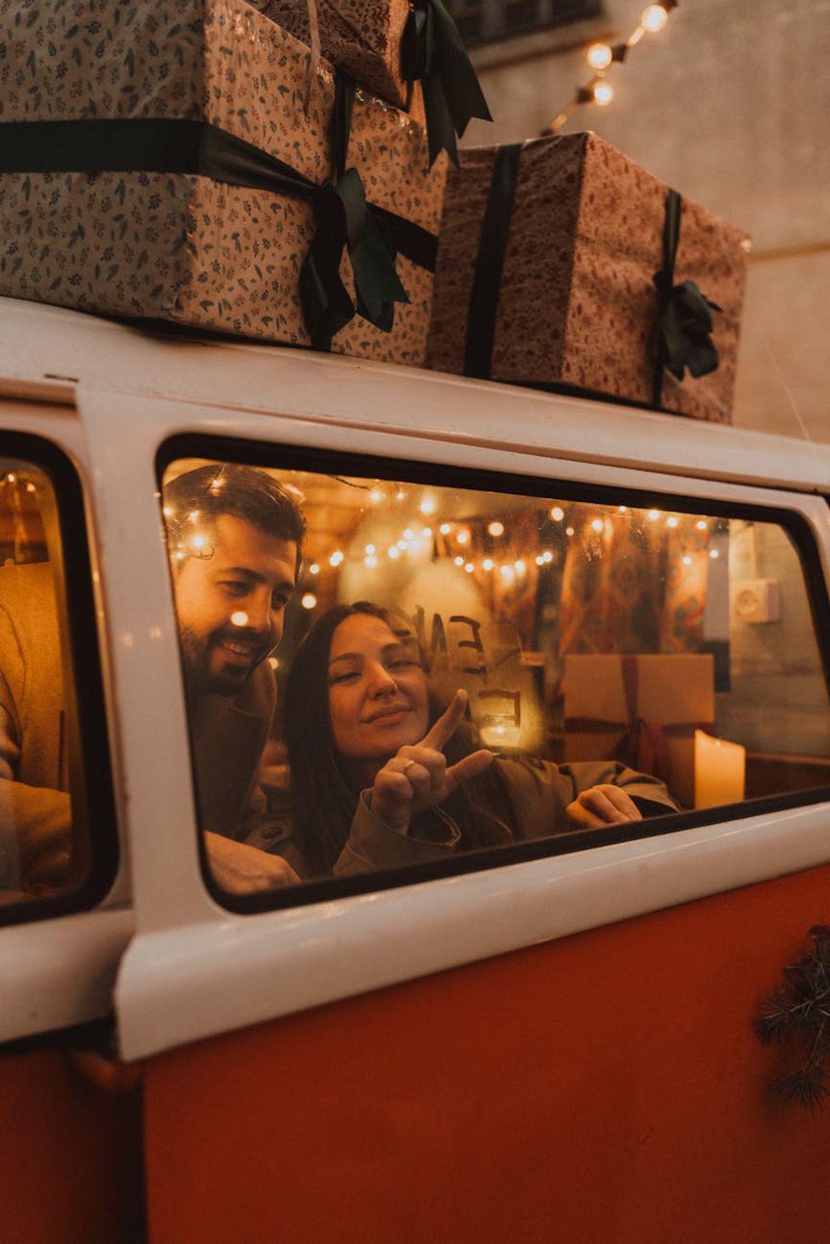 A joyful couple in a decorated van surrounded by festive holiday lights and stacked gifts, embodying holiday spirit.