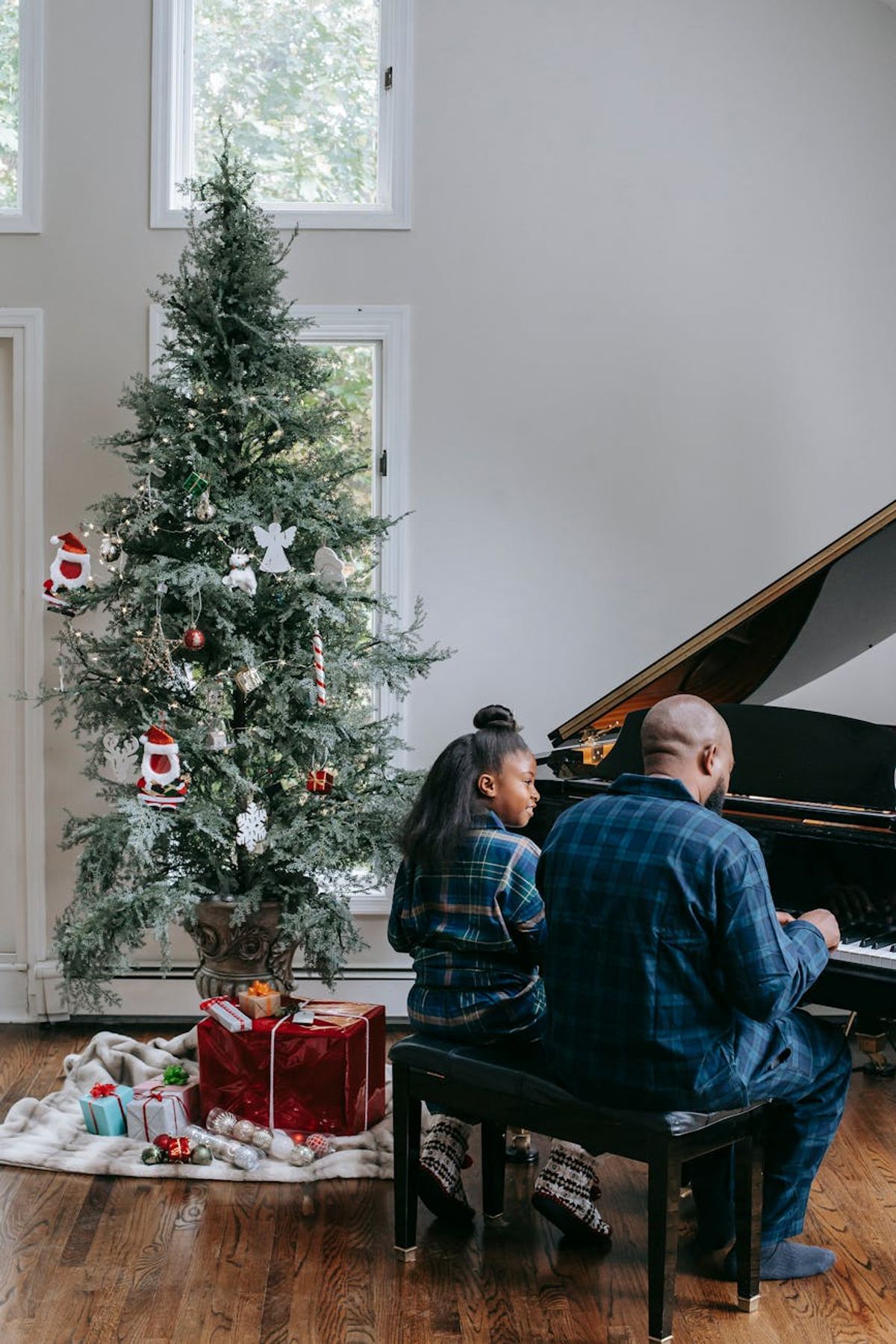 A father and daughter enjoy playing piano together beside a decorated Christmas tree indoors.