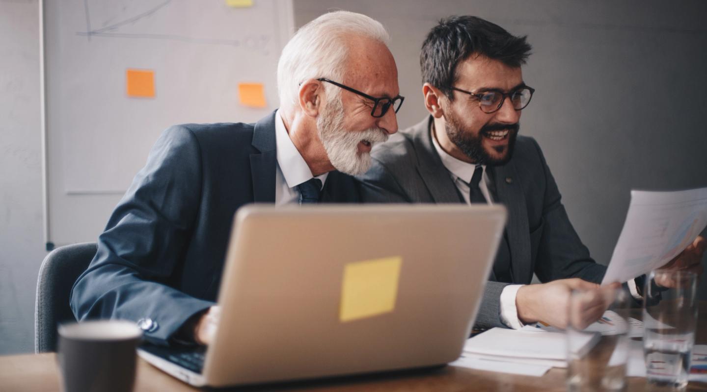 Due professionisti, uno senior con barba bianca e uno più giovane, esaminano documenti davanti a un laptop in ufficio.