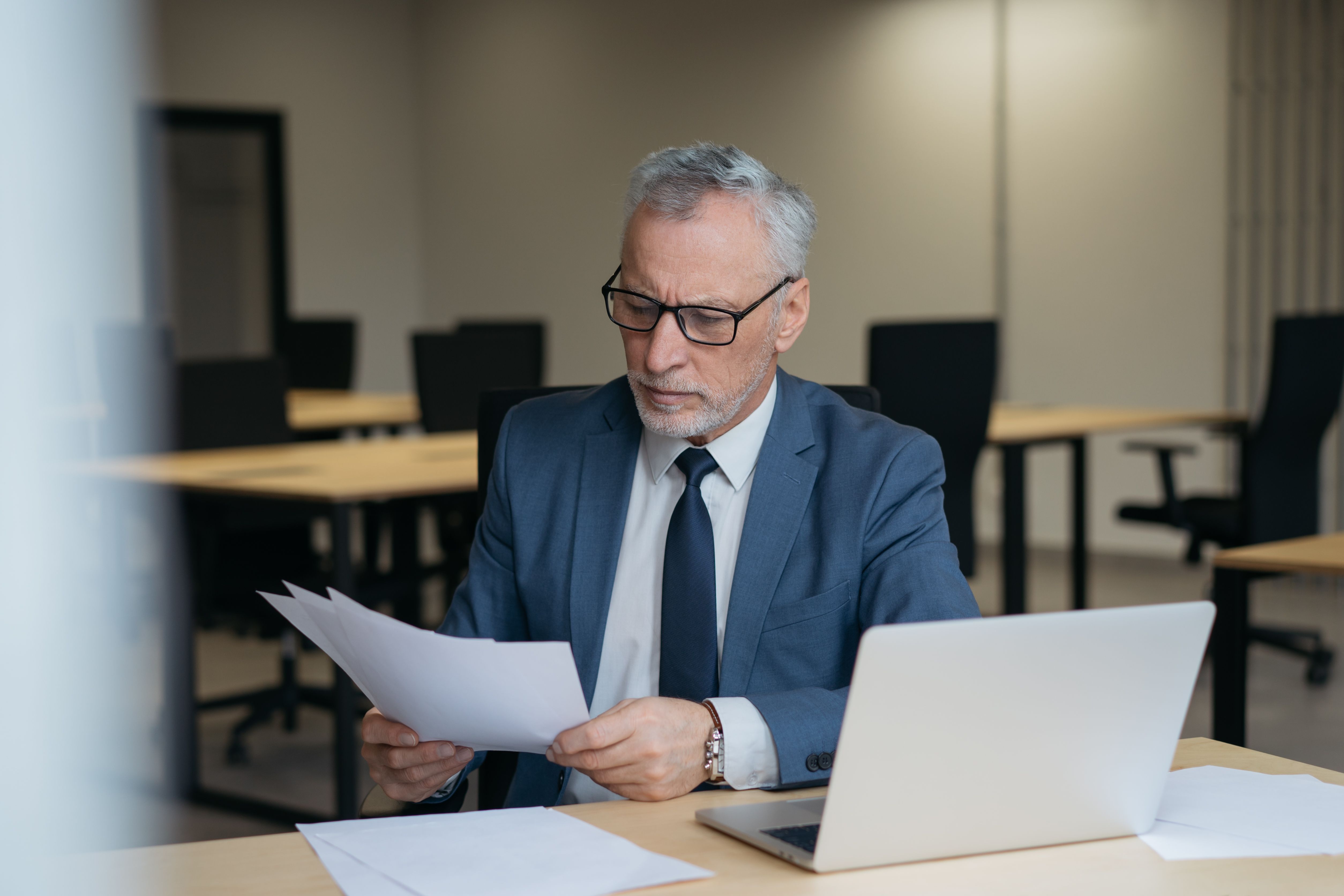 Un uomo con cappelli grigi e occhiali seduto a una scrivania in un ufficio, concentrato mentre legge dei documenti cartacei. 