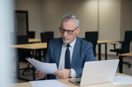 Un uomo con cappelli grigi e occhiali seduto a una scrivania in un ufficio, concentrato mentre legge dei documenti cartacei.