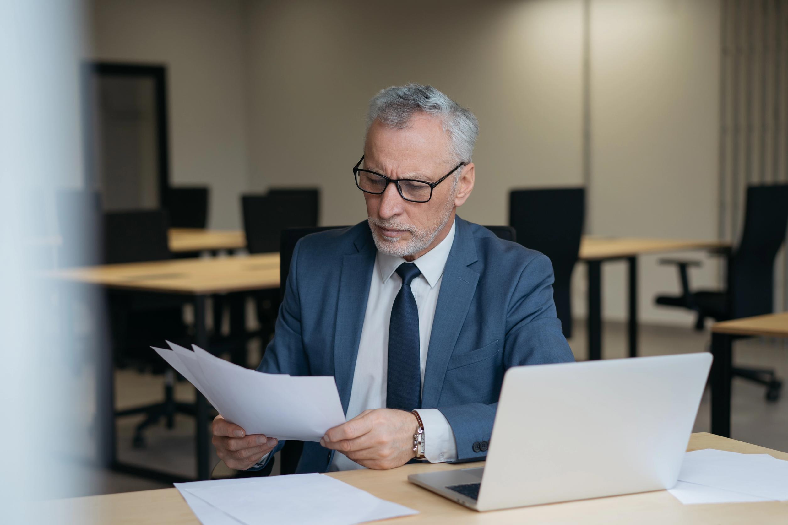 Un uomo con cappelli grigi e occhiali seduto a una scrivania in un ufficio, concentrato mentre legge dei documenti cartacei.