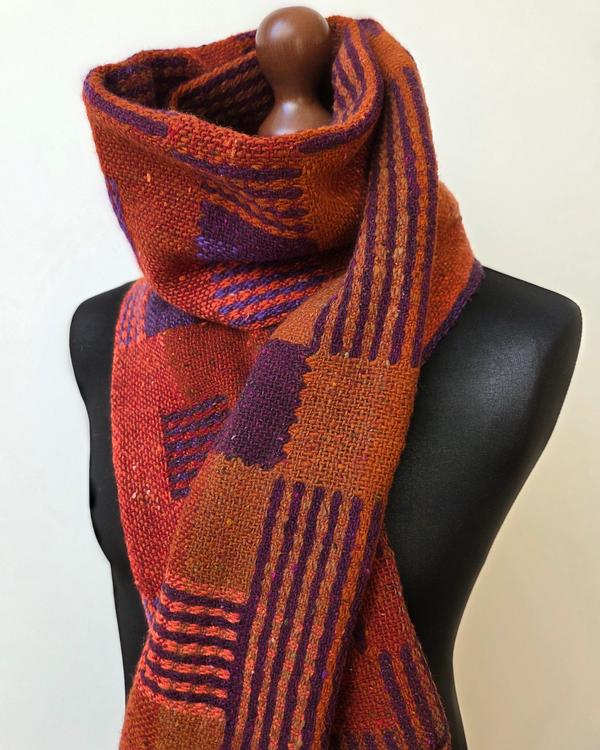 Handwoven merino wool scarf in shades of orange, red, violet and purple, draped on a mannequin. The geometric pattern consists of alternating colour fields and bands.