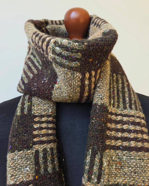 Handwoven artisan scarf made from Irish Donegal merino wool, displayed on a black mannequin. A geometric pattern in natural beige, olive green and deep brown highlights the craftsmanship and timeless character of the woven textile.