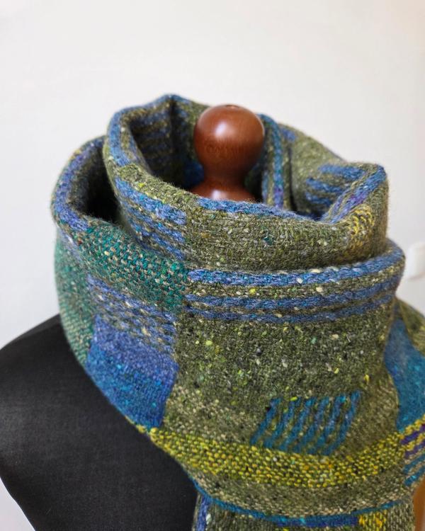 Close-up of a handwoven wool scarf in shades of green, blue and turquoise, showing the soft textile texture and geometric pattern accents.