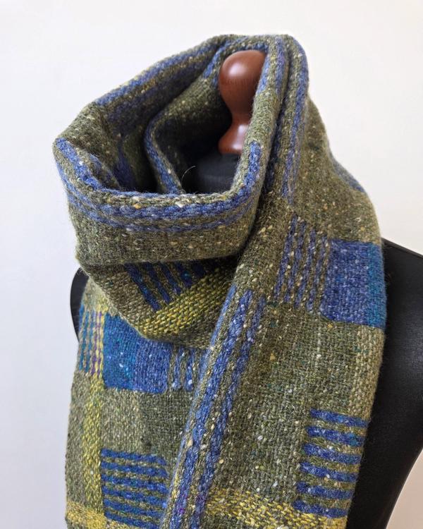Handwoven merino wool and mohair scarf in olive green and blue, draped on a mannequin, with contrasting stripes and geometric accents.