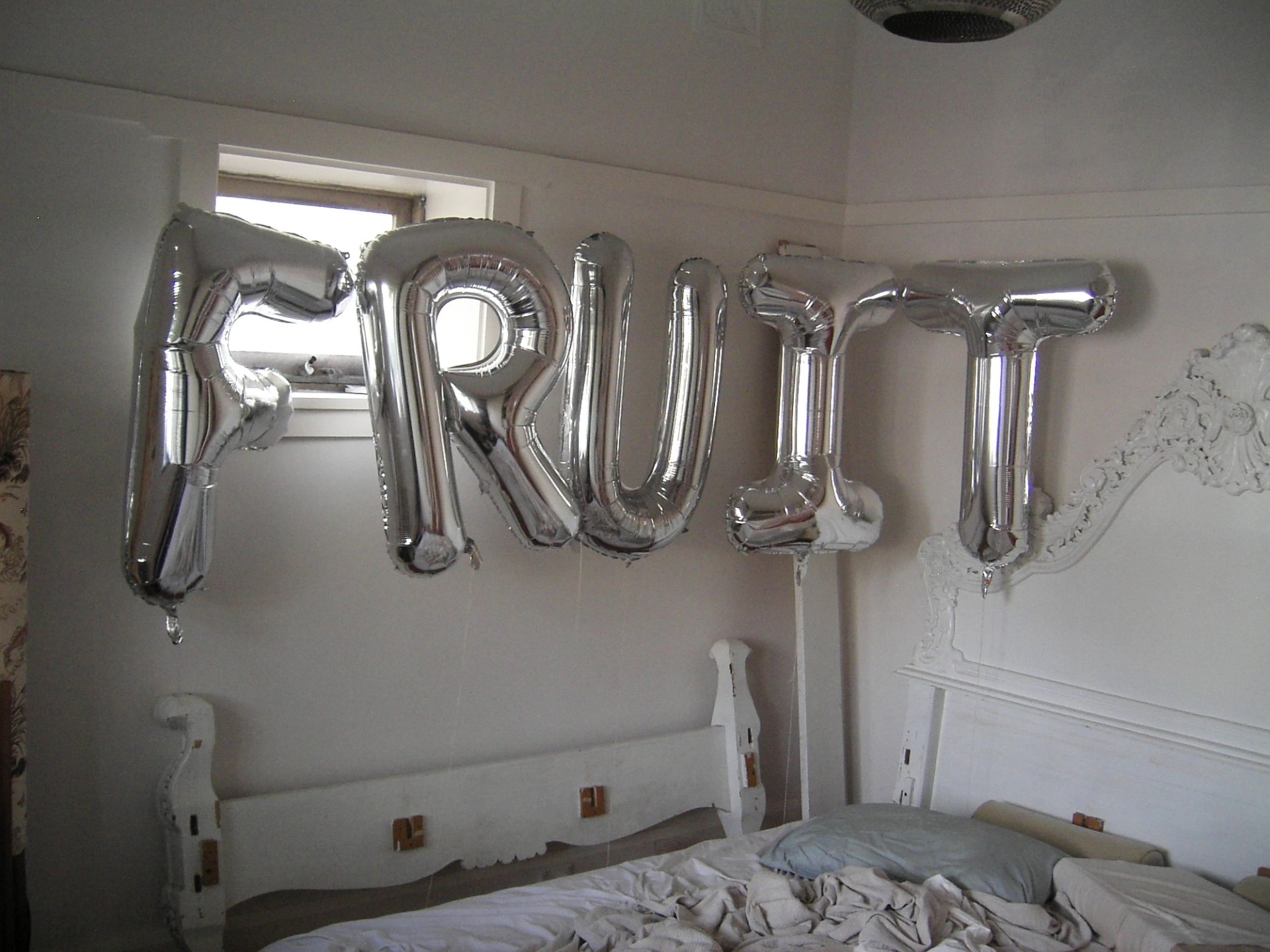 Silver balloon letters spelling out "FRUIT" in a bedroom with an unmade bed and ornate white headboard.