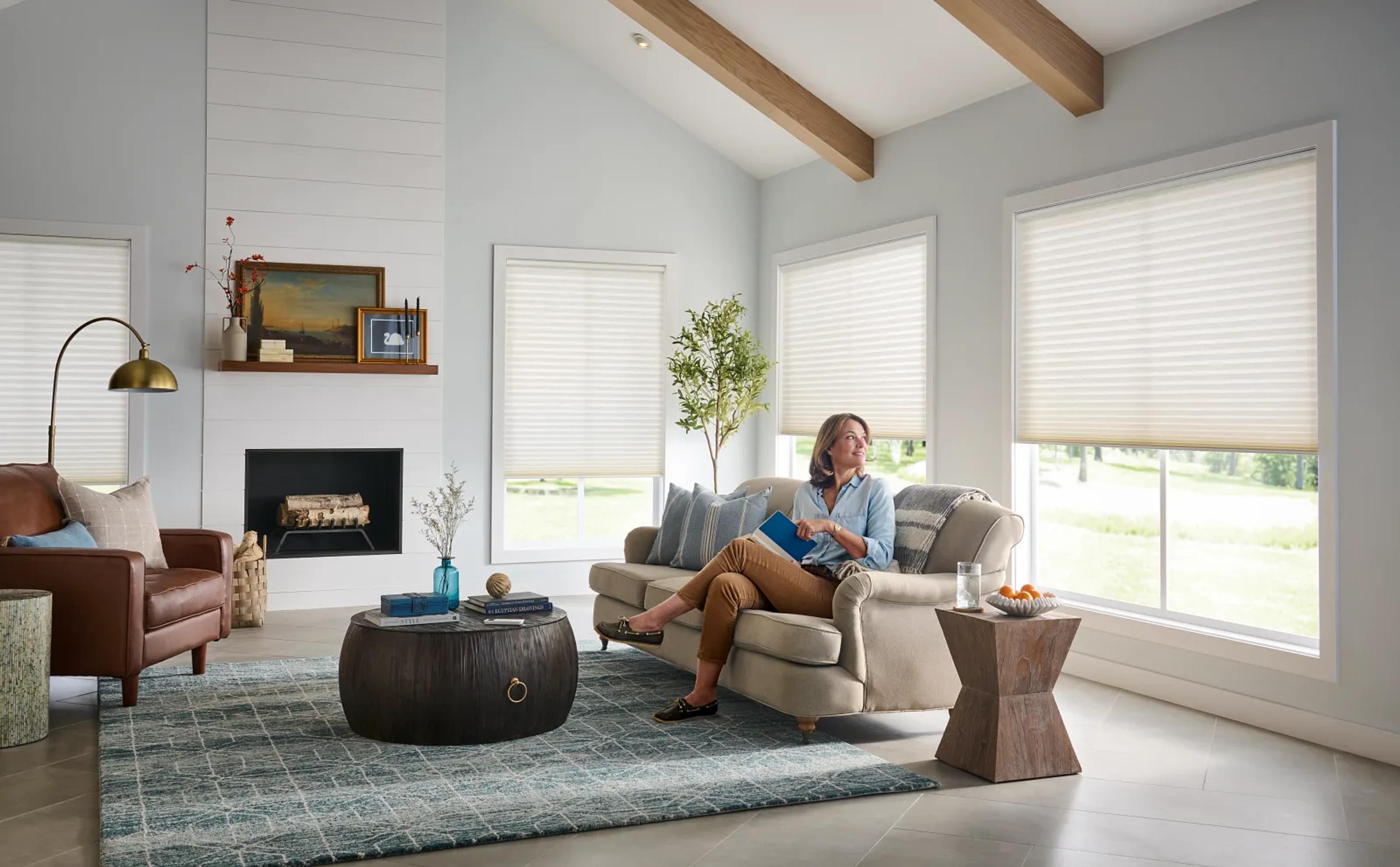 Woman sitting in a living room, reading a book, and looking out a window with Honeycomb window coverings.