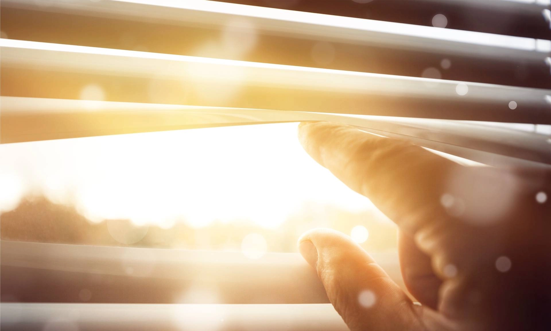 A person's hand peering through the metal window blinds as the sun streams inside