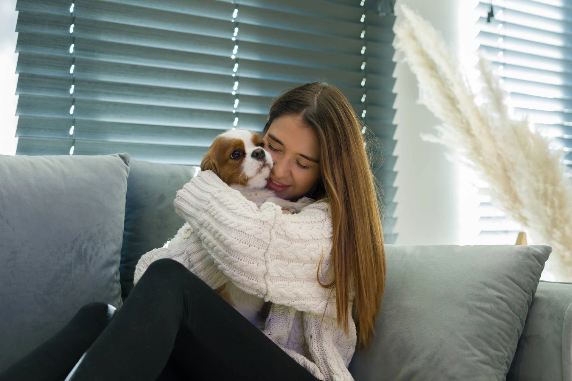 Woman holding small white dog on sofa in front of custom window blinds.