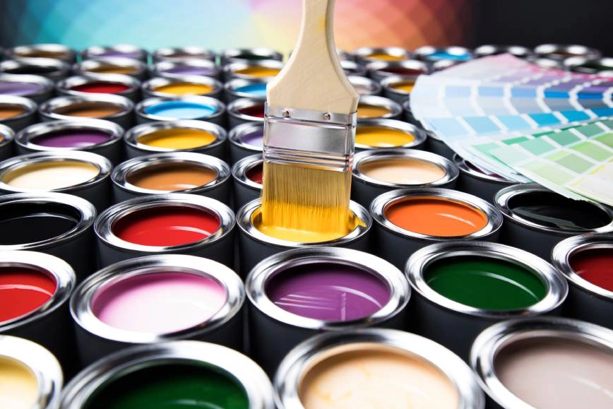 Dozens of cans of paint with a paint brush being dipped into a can of yellow paint.