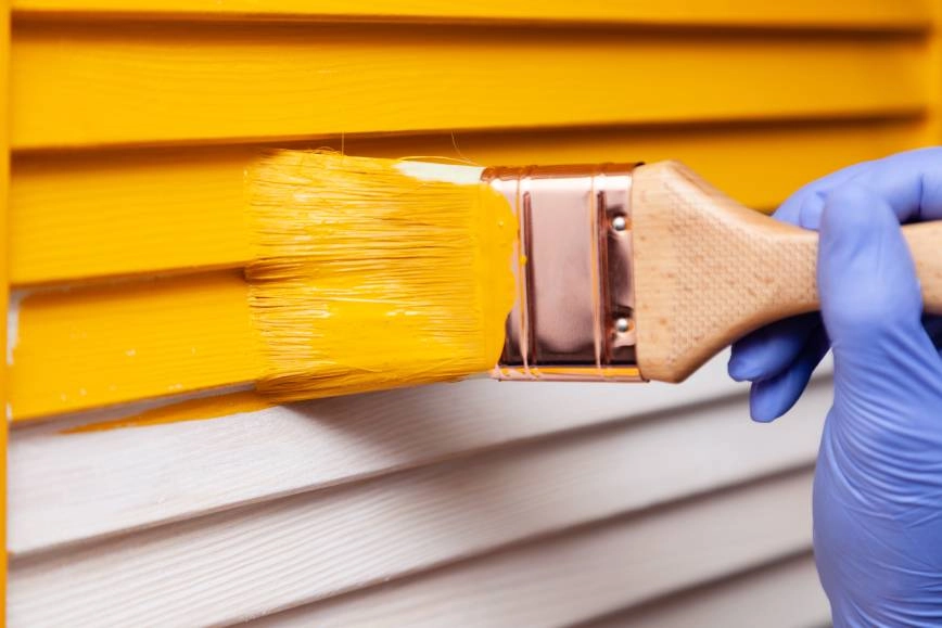 A gloved hand painting the side of a house with yellow paint.