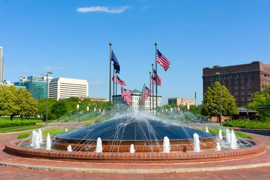 An image of a fountain and several American flags in Omaha, NE, with blue skies