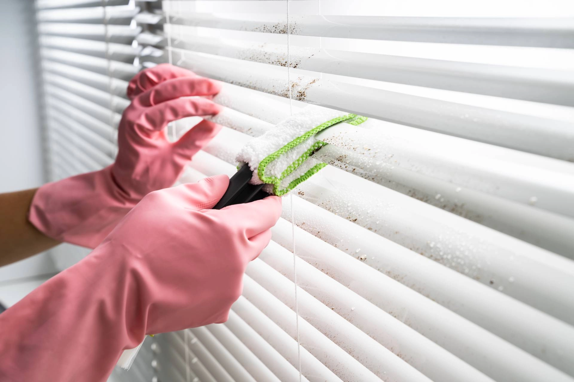 Person wearing pink gloves cleaning window blinds with a cleaning tool.