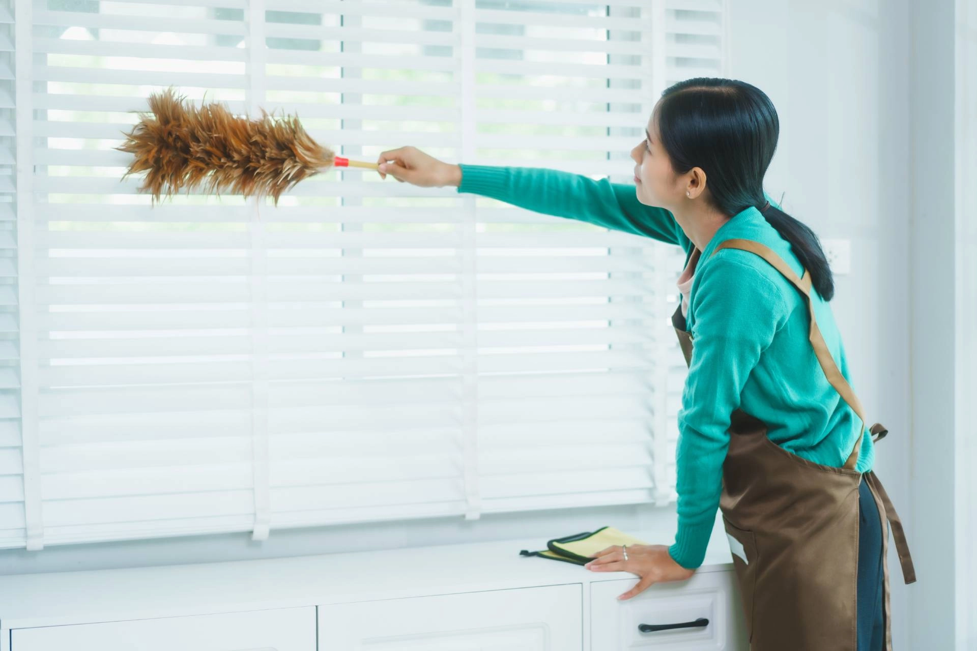 Woman in a green sweater dusting blinds with a feather duster near Brunswick, GA