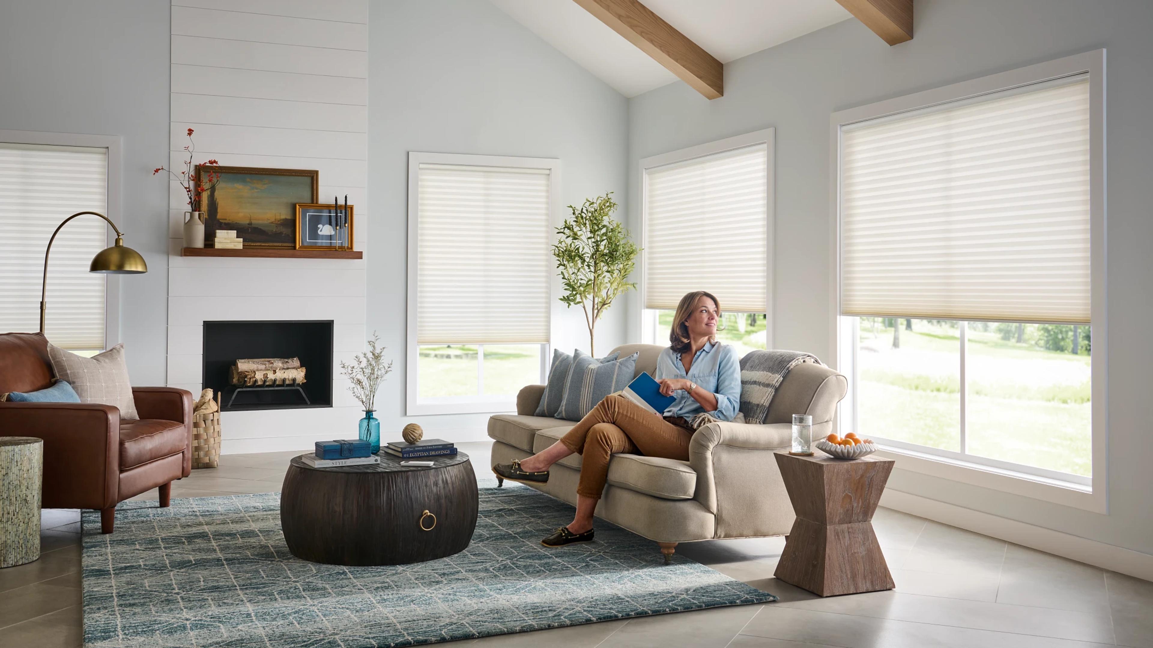 Woman sitting in a living room, reading a book, and looking out a window with Honeycomb window coverings.