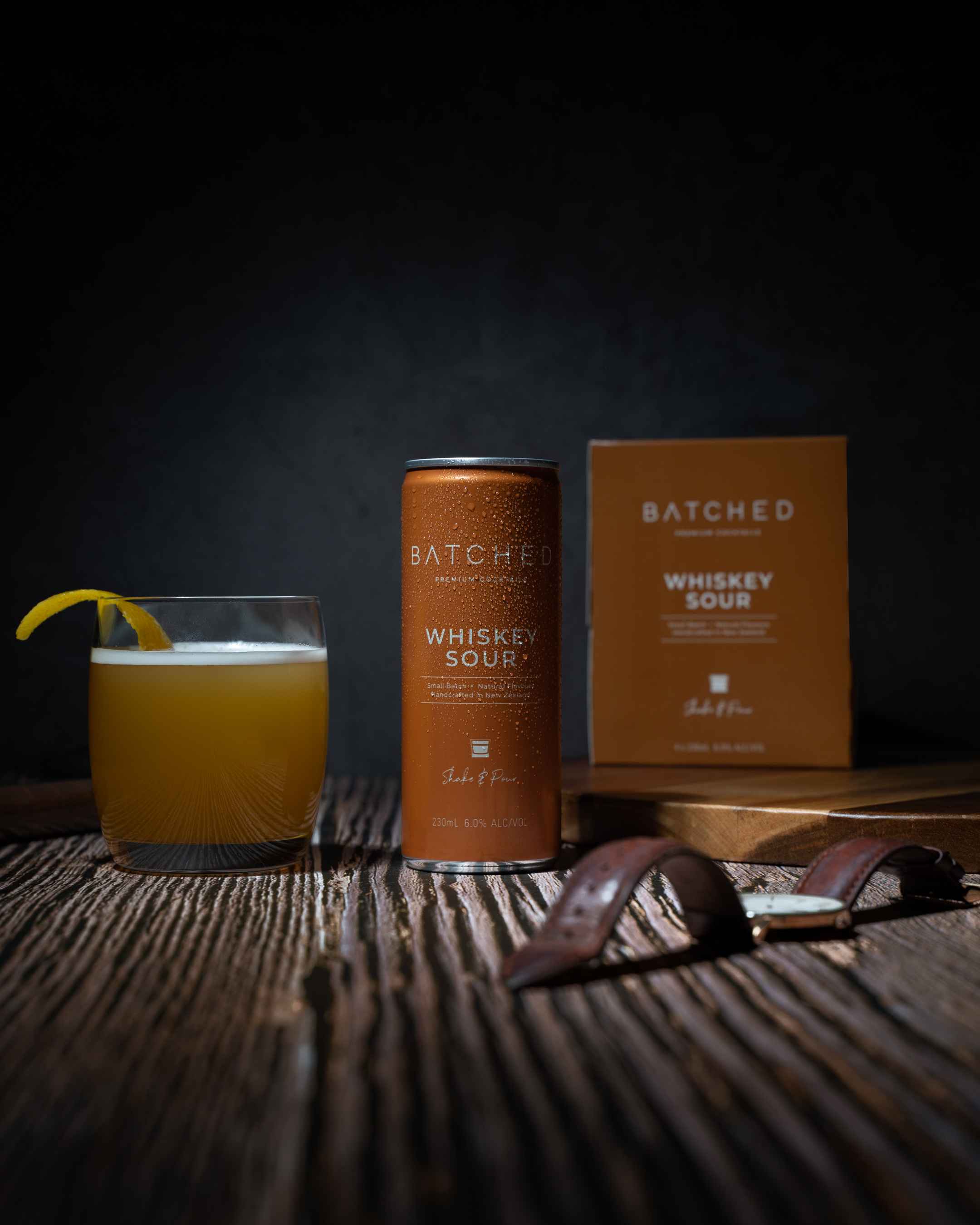 Can, box and glass of Batched Whiskey Sour on wooden table