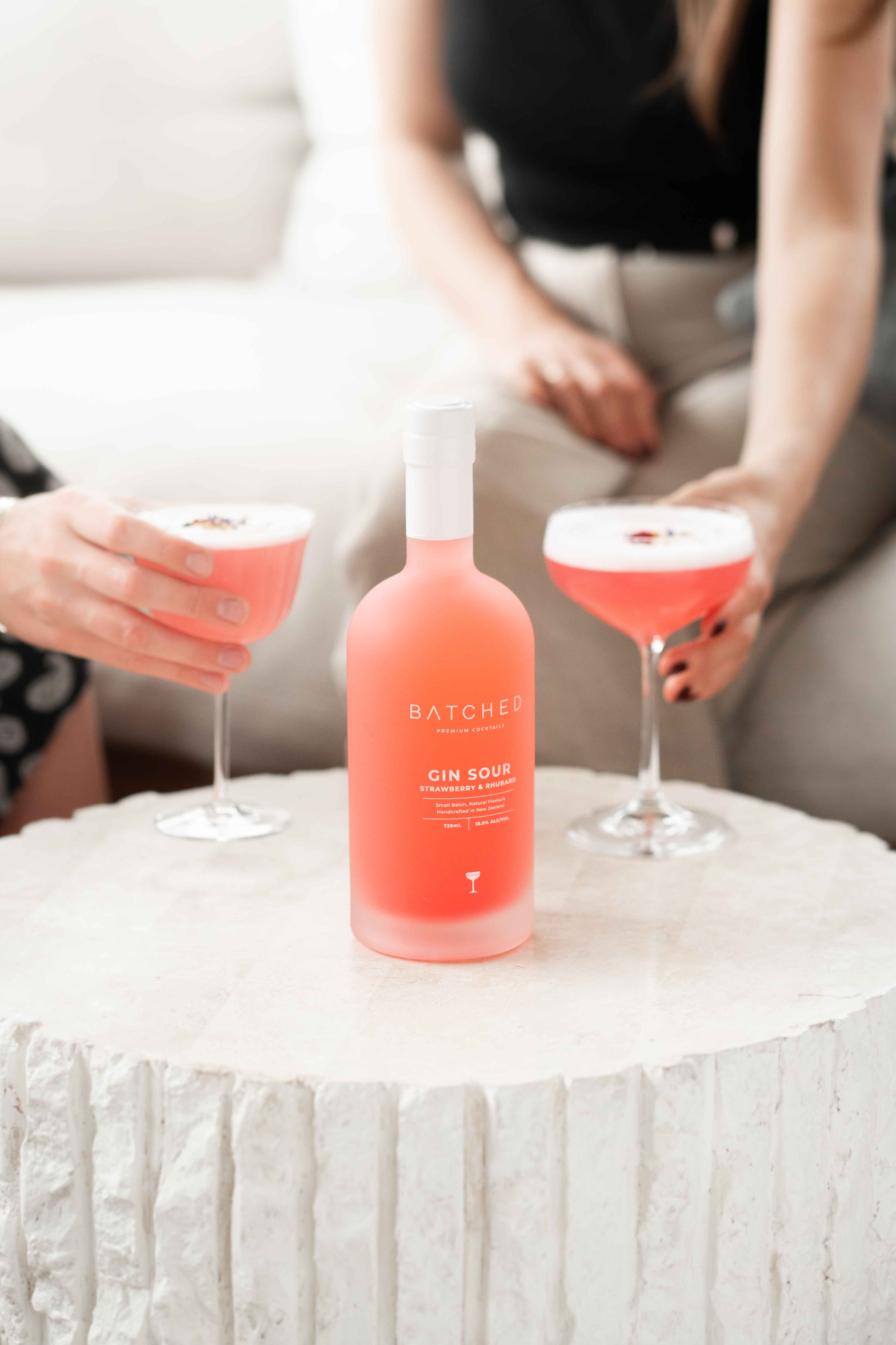 Batched Gin Sour bottle on table with two people holding glasses in the background