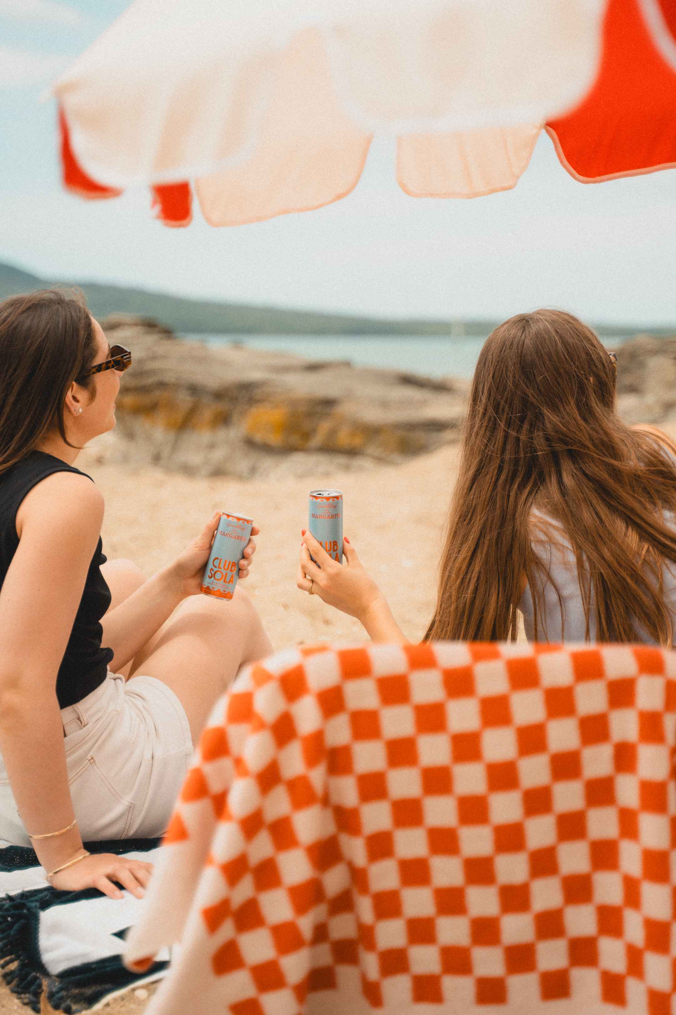 Two girls drinking cans of Club Sola while sitting on the beach