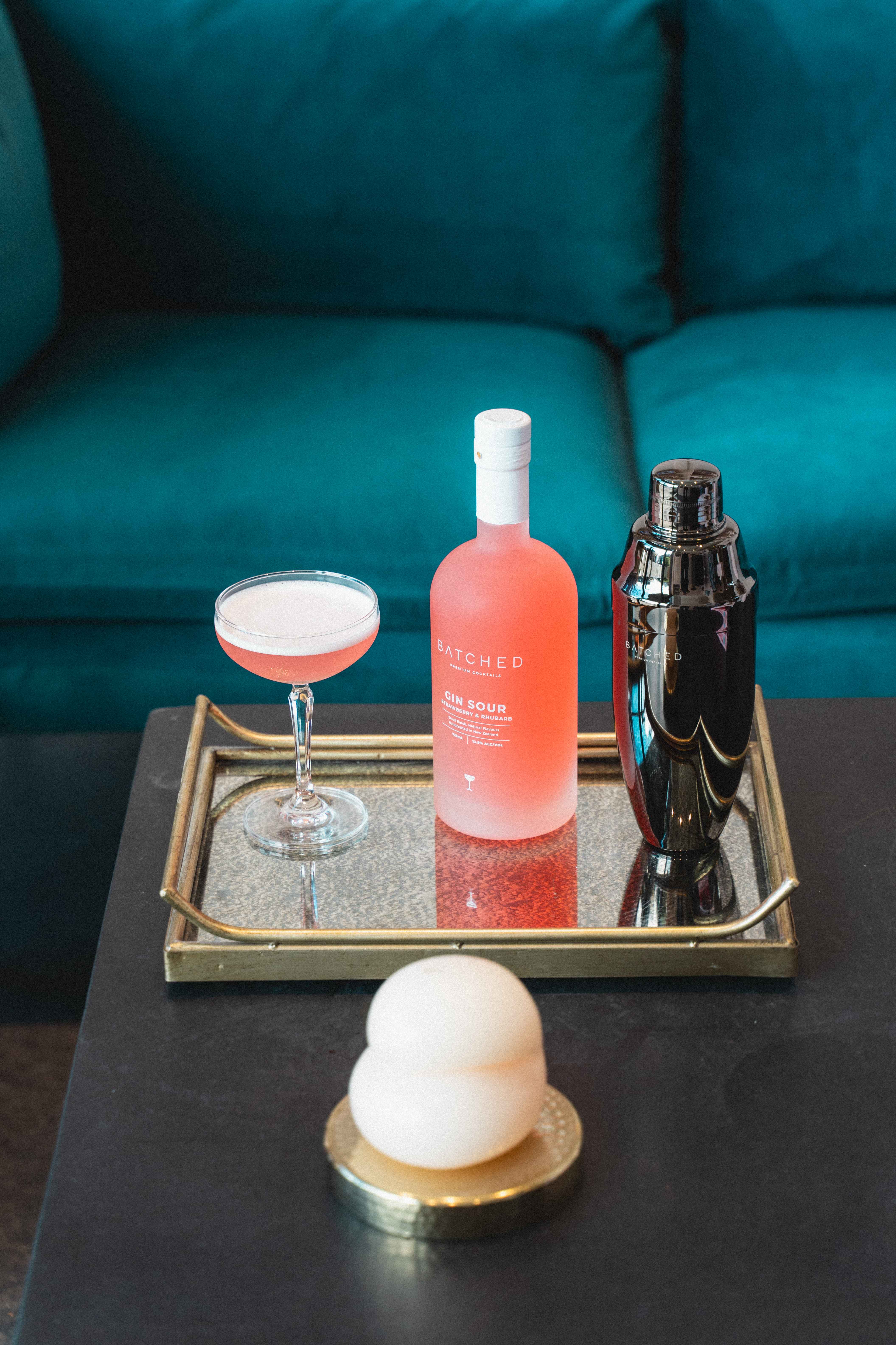 Batched Gin Sour bottle, glass and shaker sitting on a tray with couch in background