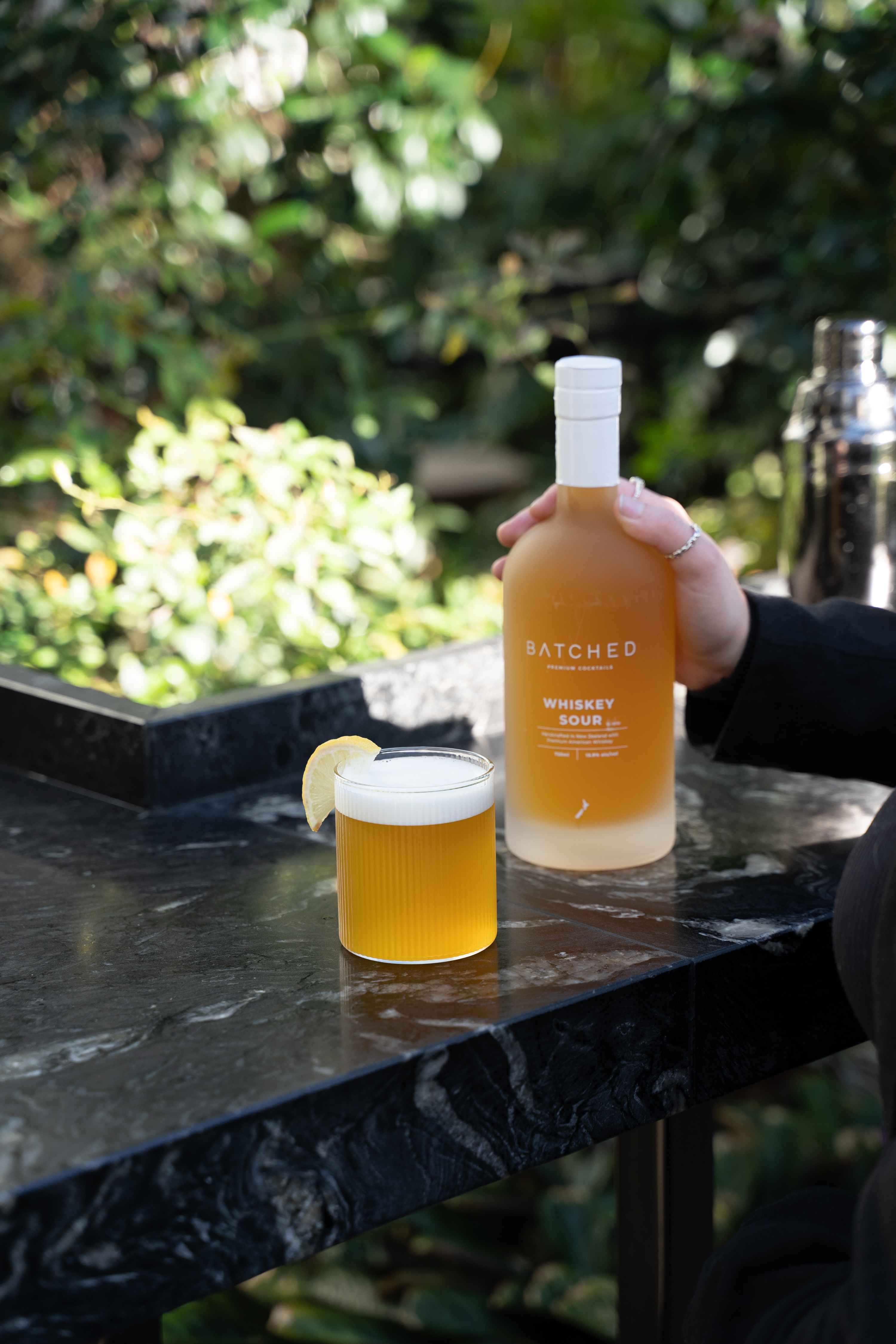 Batched Whiskey Sour bottle with drink served in glass being held on table