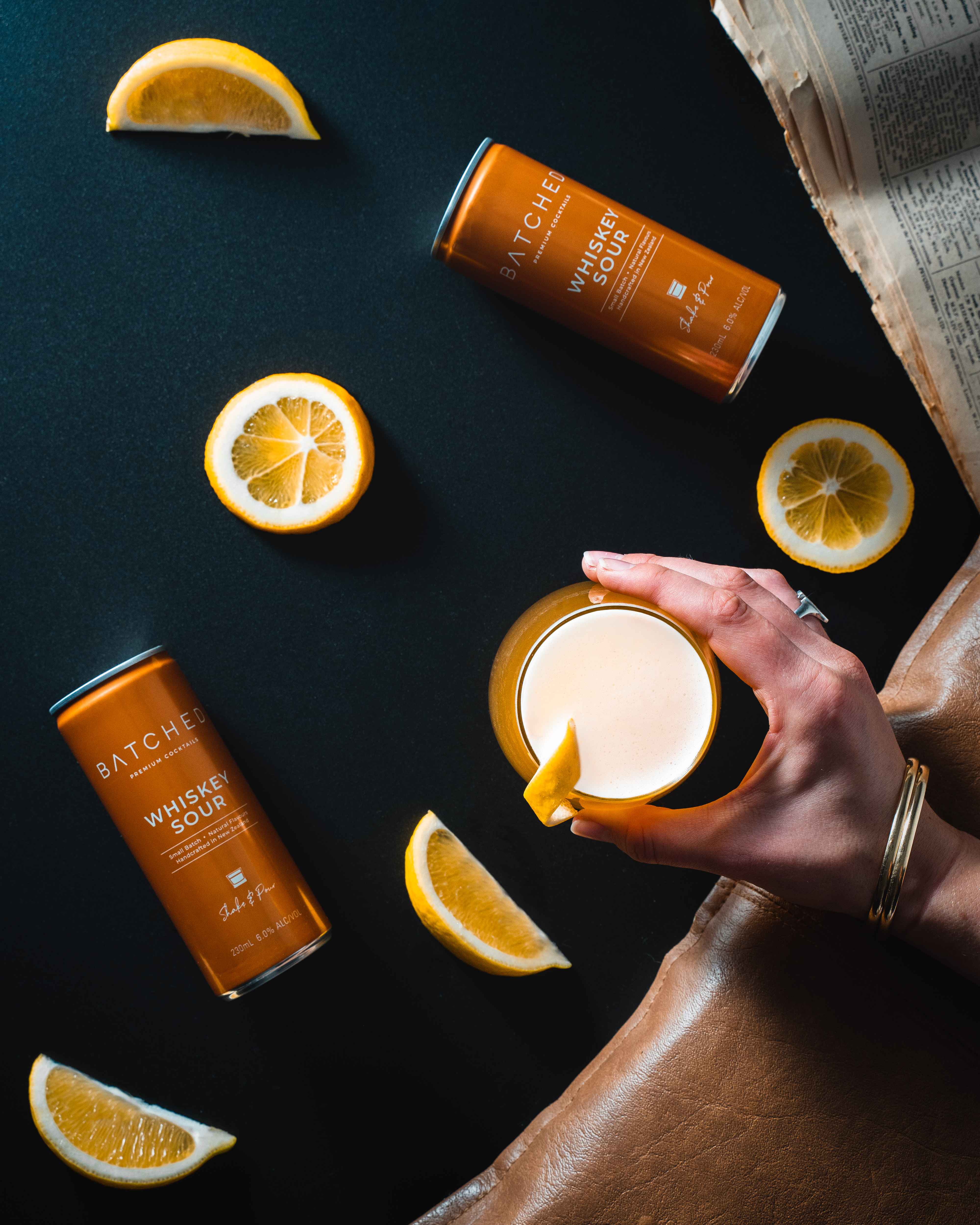 Top view of Batched Whiskey Sour cans, served in a glass held by a hand, with lemon slices on a table