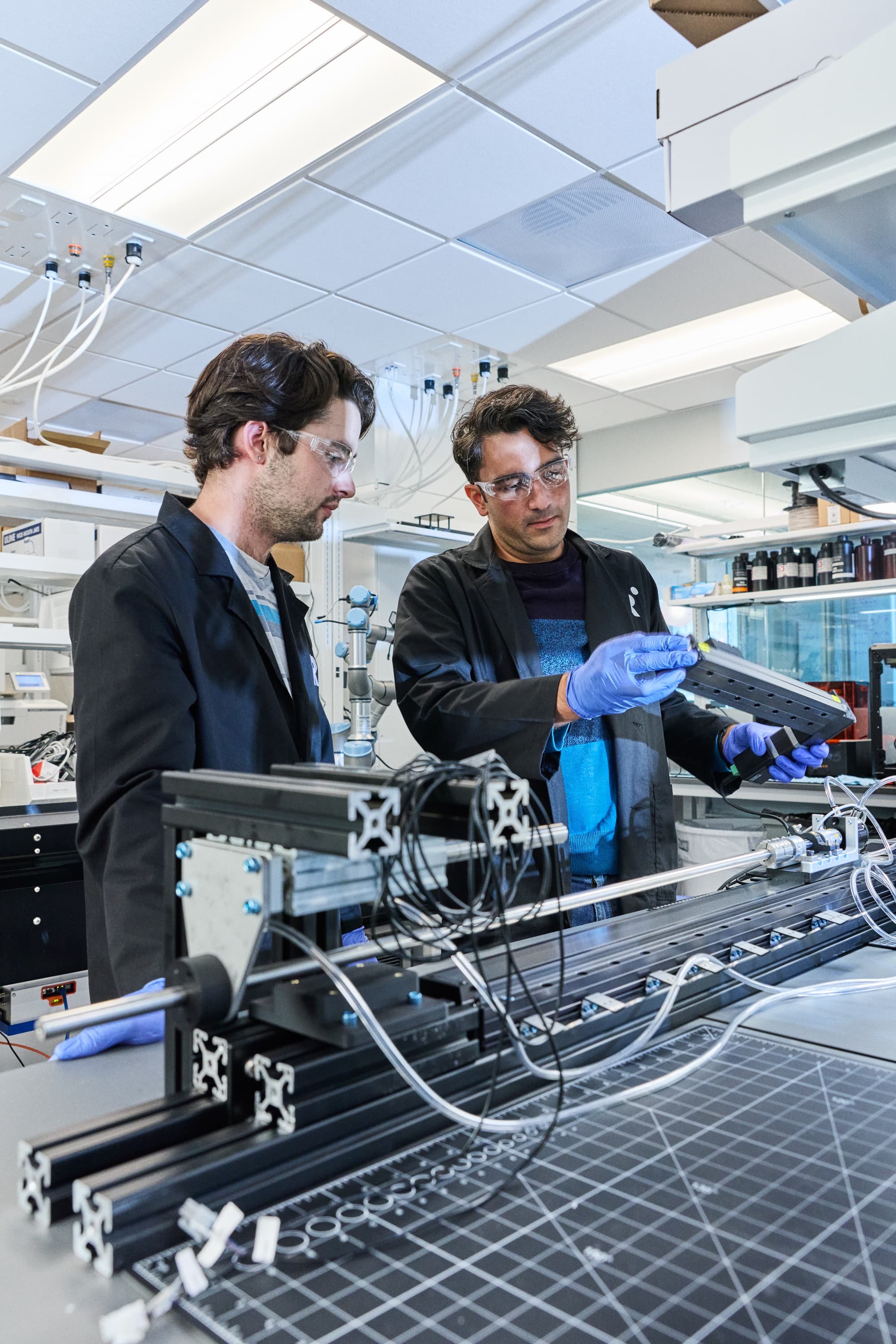 Two people wearing black lab coats and blue gloves working with a robot